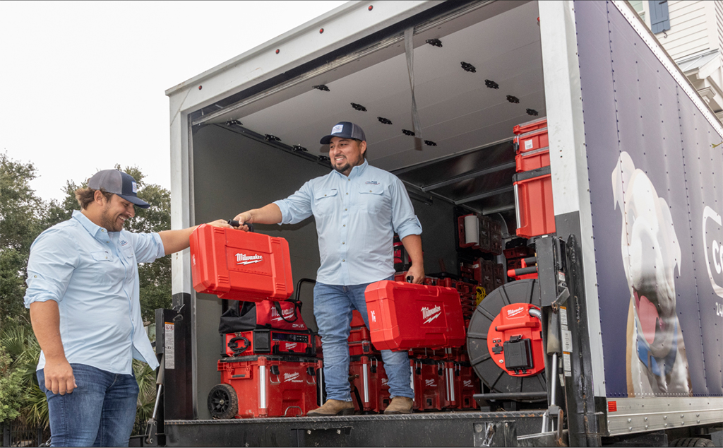 Two Blue Collars technicians handing a red tool case between them at the open back of the company box truck, loaded with tool cases and a drain cable reel
